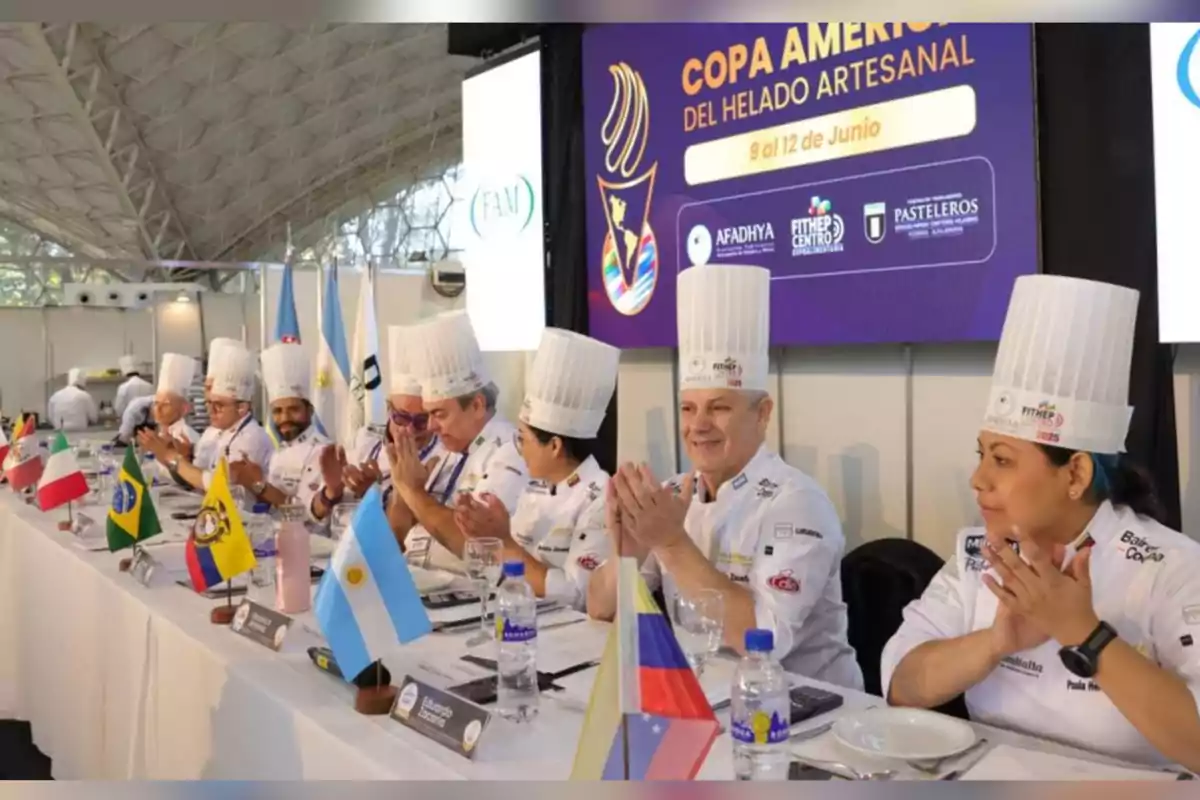 Un grupo de jueces con gorros de chef sentados en una mesa larga, aplaudiendo durante un evento de la Copa América del Helado Artesanal, con banderas de varios países y un cartel del evento al fondo. Un grupo de jueces con gorros de chef sentados en una mesa larga, aplaudiendo durante un evento de la Copa América del Helado Artesanal, con banderas de varios países y un cartel del evento al fondo.