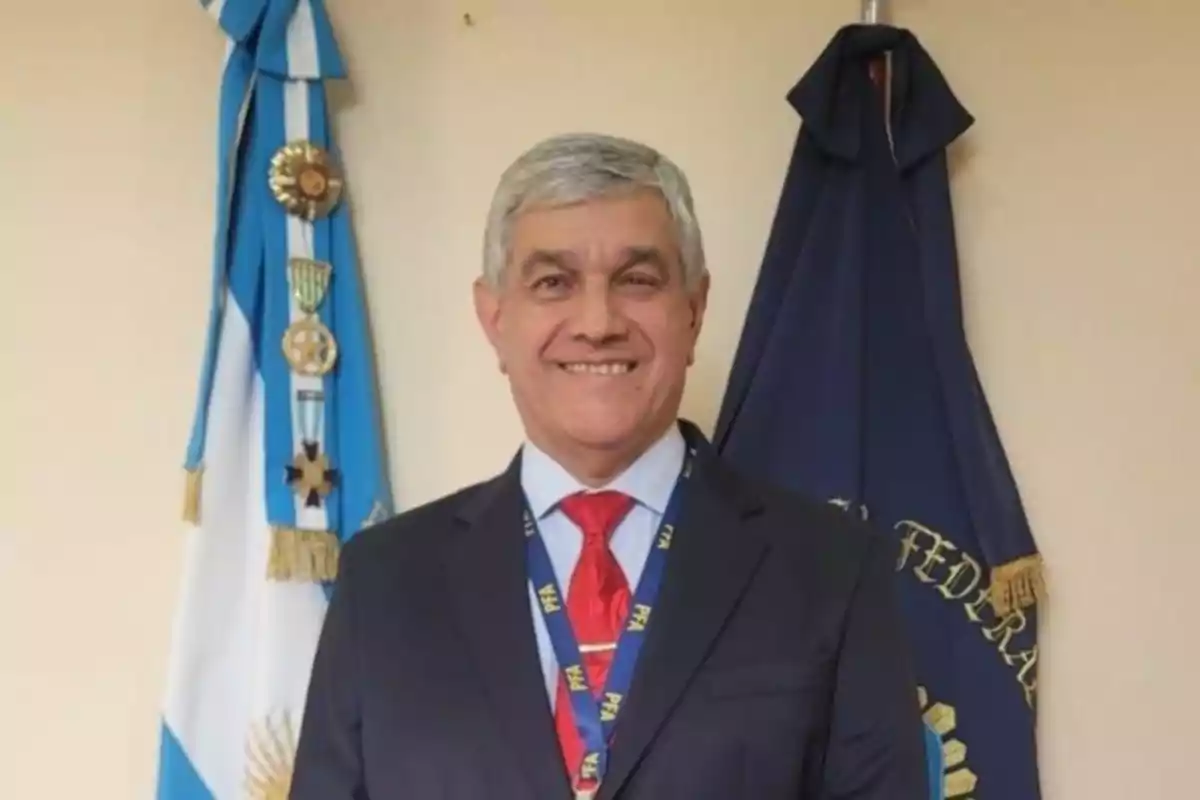 Hombre de cabello canoso con traje y corbata roja sonriendo frente a banderas oficiales