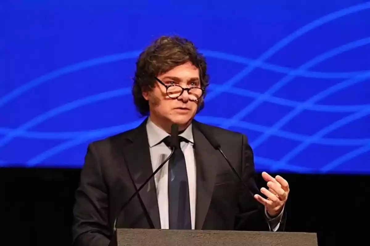 Hombre de cabello rizado y gafas hablando en un podio con fondo azul geométrico