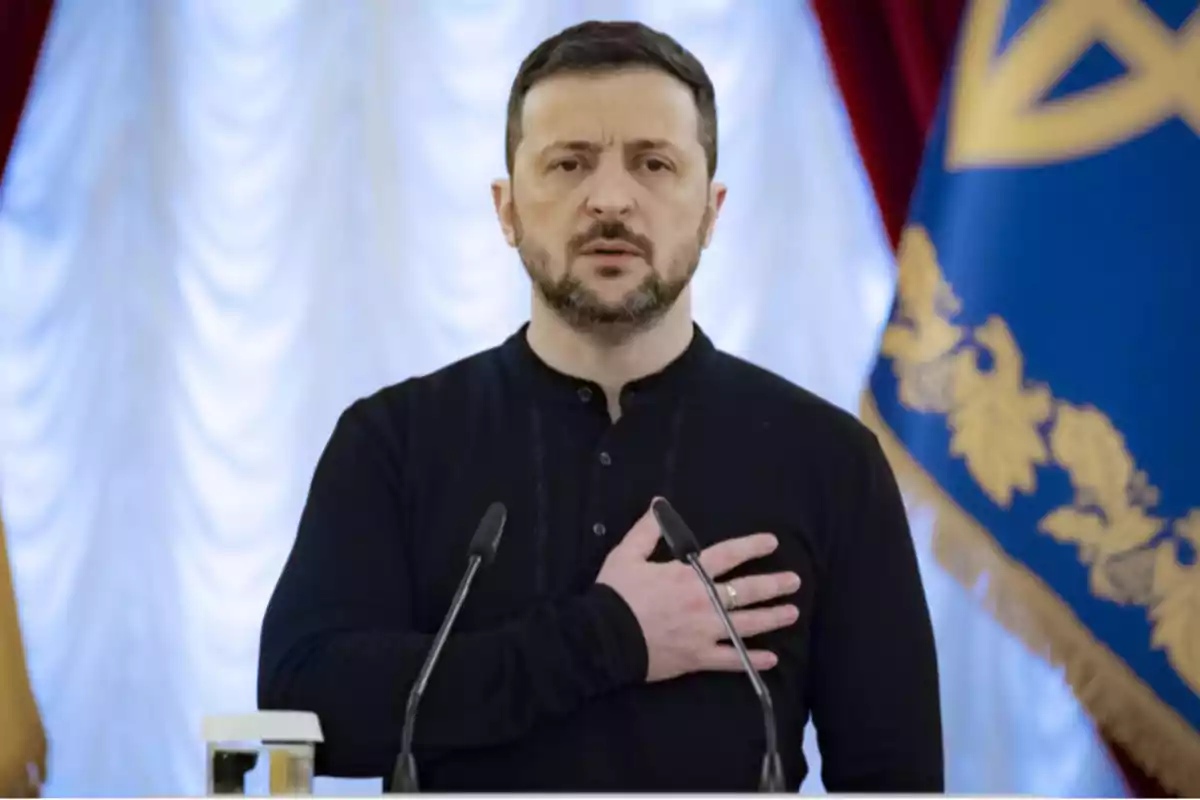 Un hombre con barba y cabello corto, vestido de negro, con la mano en el pecho, frente a dos micrófonos y un fondo con cortinas blancas y una bandera azul con detalles dorados. Un hombre con barba y cabello corto, vestido de negro, con la mano en el pecho, frente a dos micrófonos y un fondo con cortinas blancas y una bandera azul con detalles dorados.