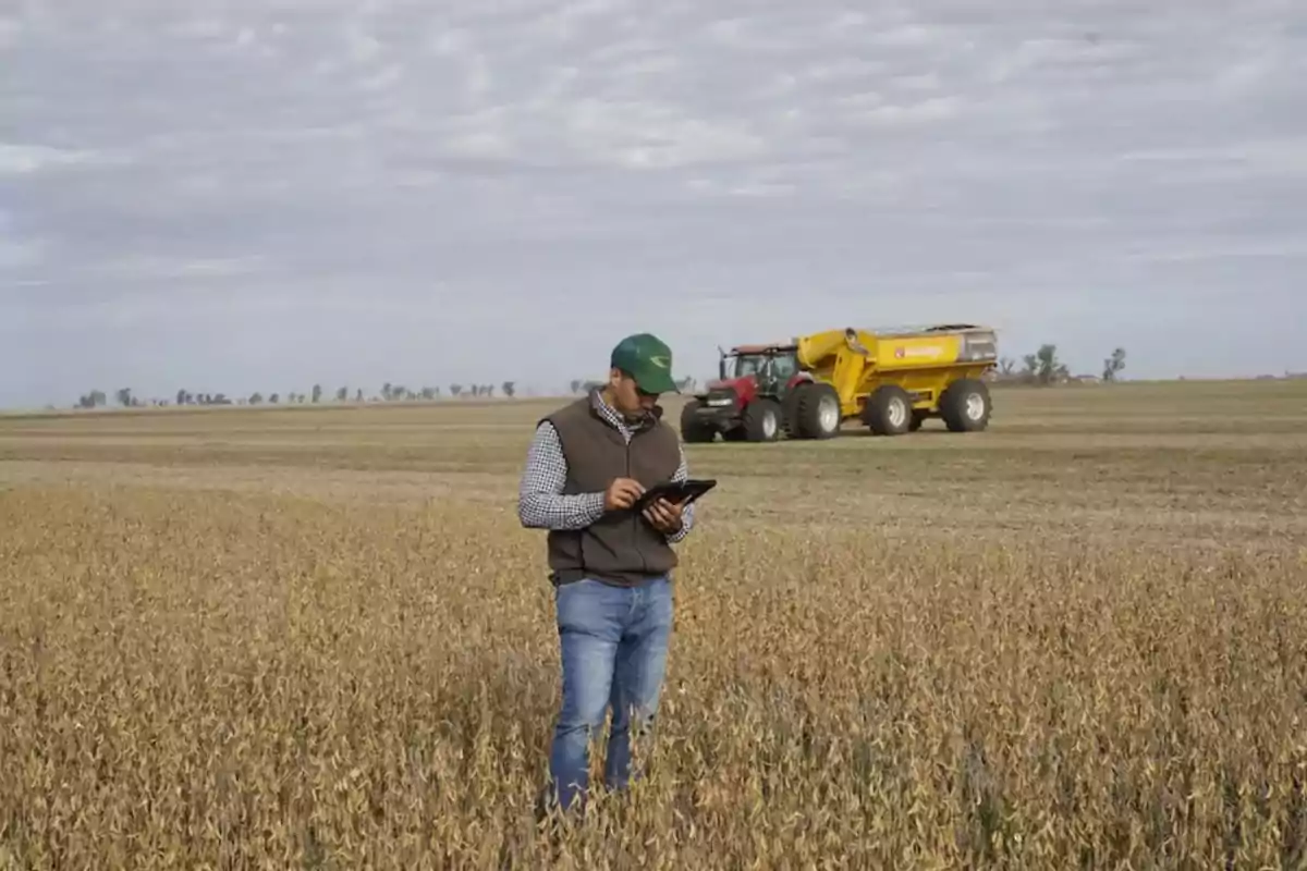 Man wearing a green cap using a tablet in a crop field with a tractor and agricultural machinery in the background
