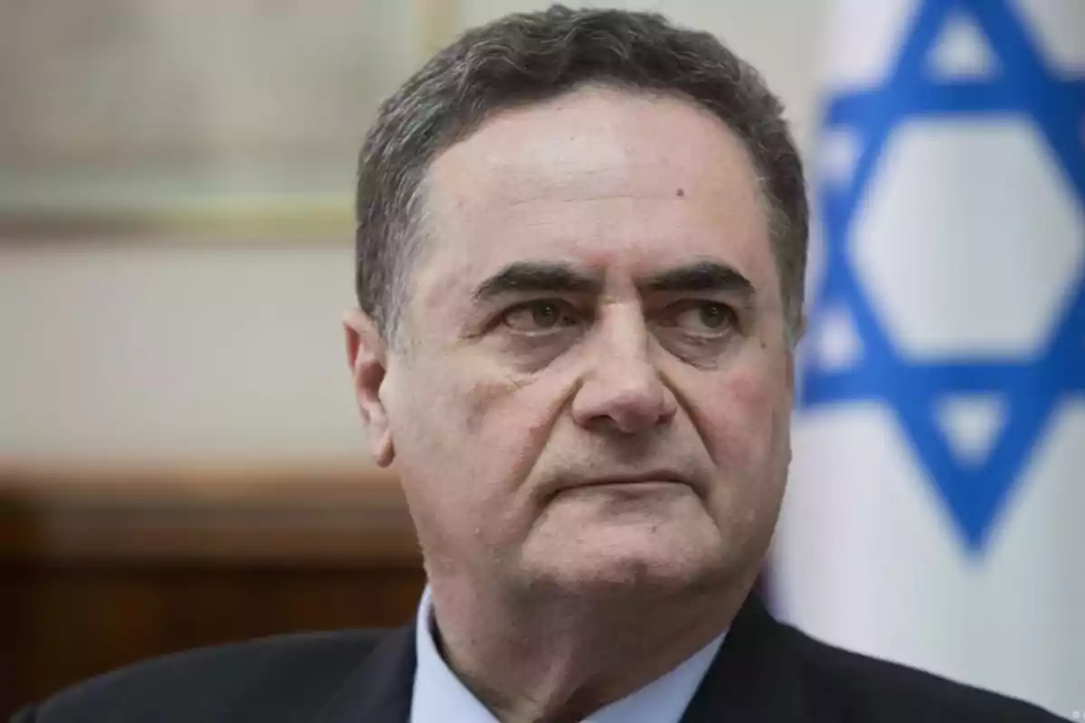 Short-haired, gray-haired man with a serious expression in front of a blurred Israeli flag in the background Short-haired, gray-haired man with a serious expression in front of a blurred Israeli flag in the background