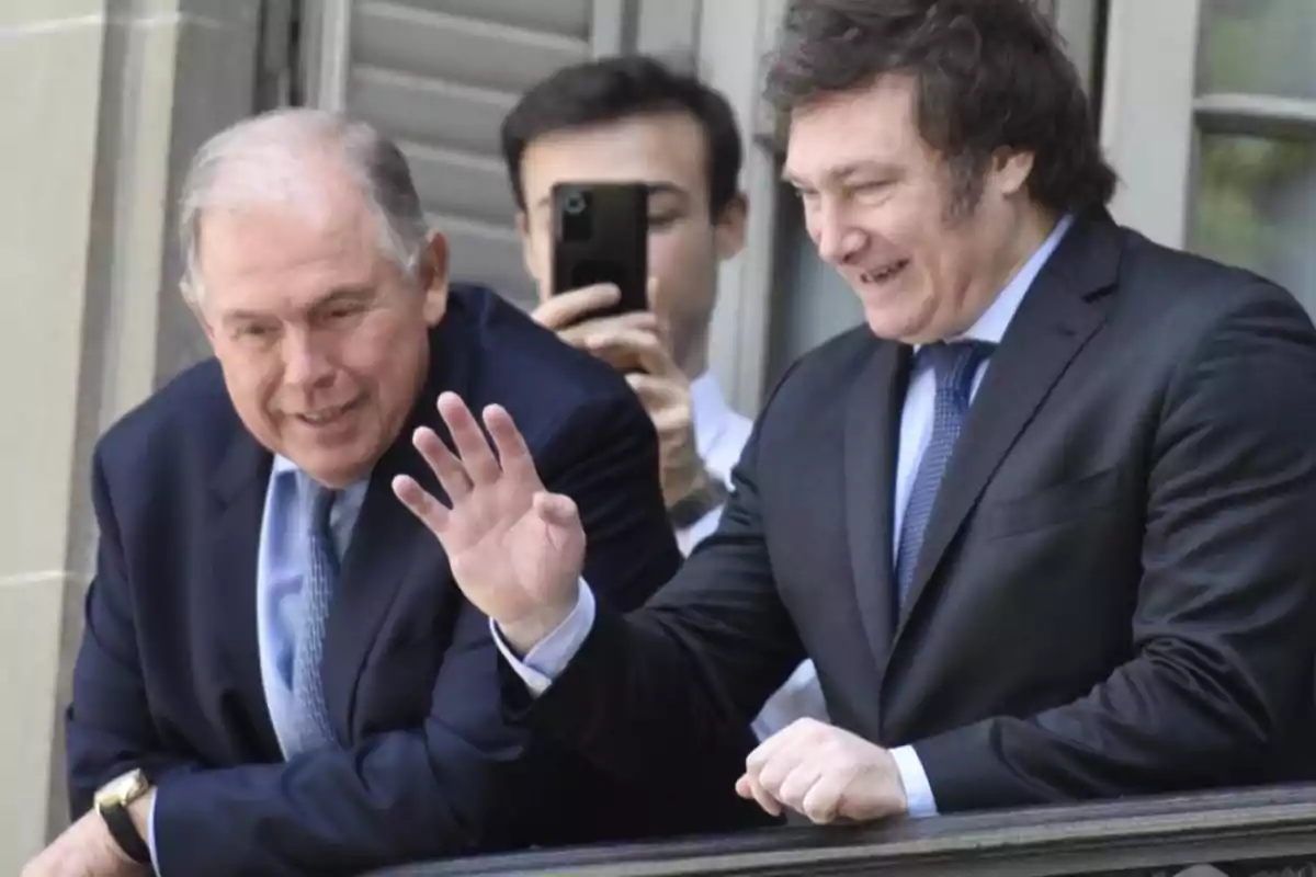 Dos hombres en traje sonríen y saludan desde un balcón mientras una persona detrás de ellos toma una foto con un teléfono móvil.