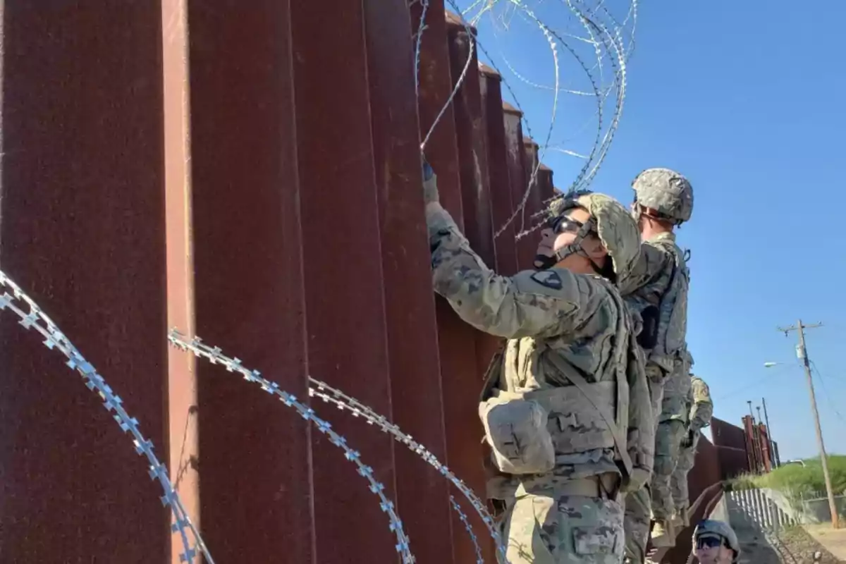 Soldados en uniforme militar instalan alambre de púas en una valla alta bajo un cielo despejado.