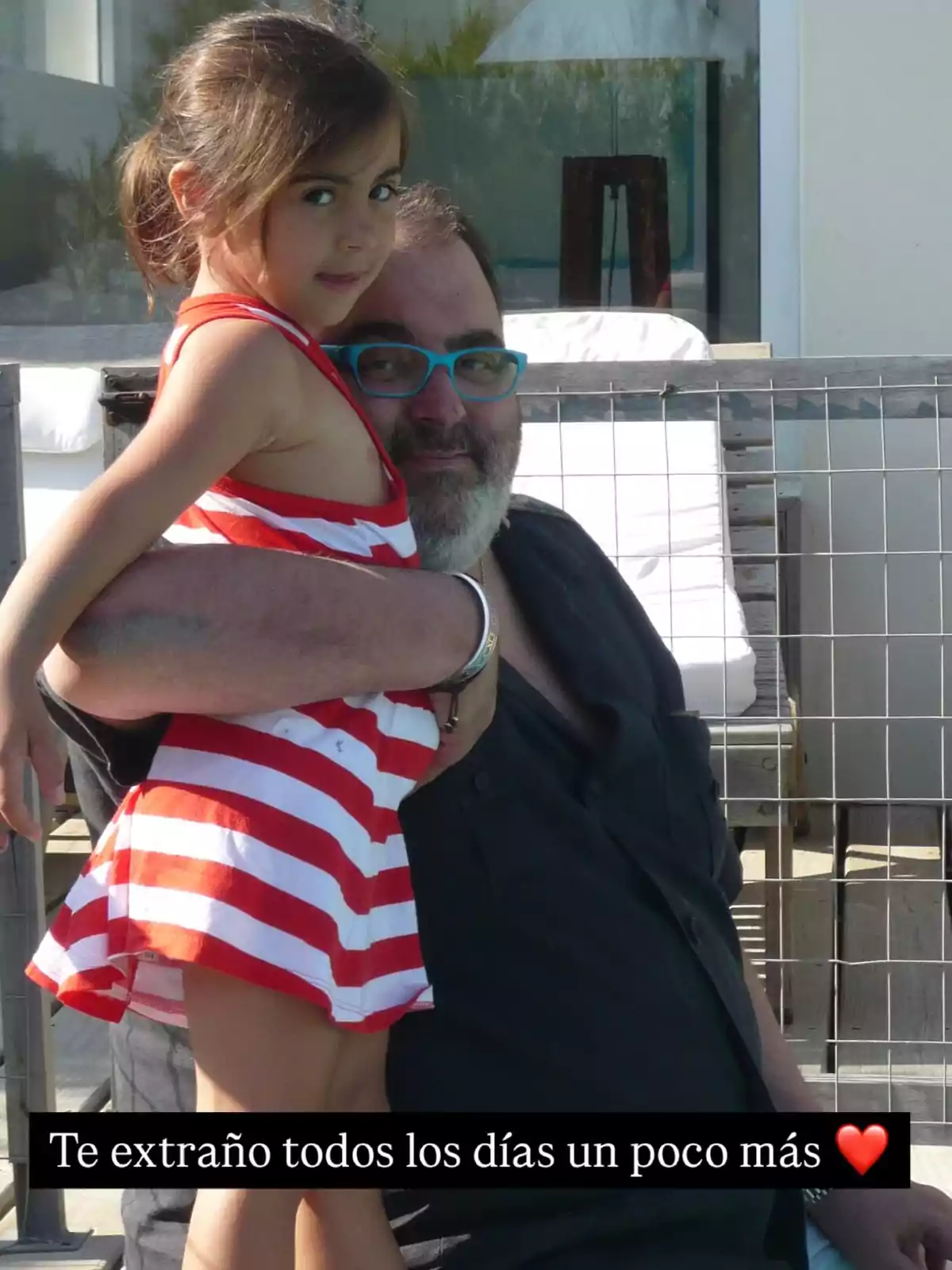 Un hombre con gafas sostiene a una niña con vestido de rayas rojas y blancas mientras están al aire libre. Un hombre con gafas sostiene a una niña con vestido de rayas rojas y blancas mientras están al aire libre.