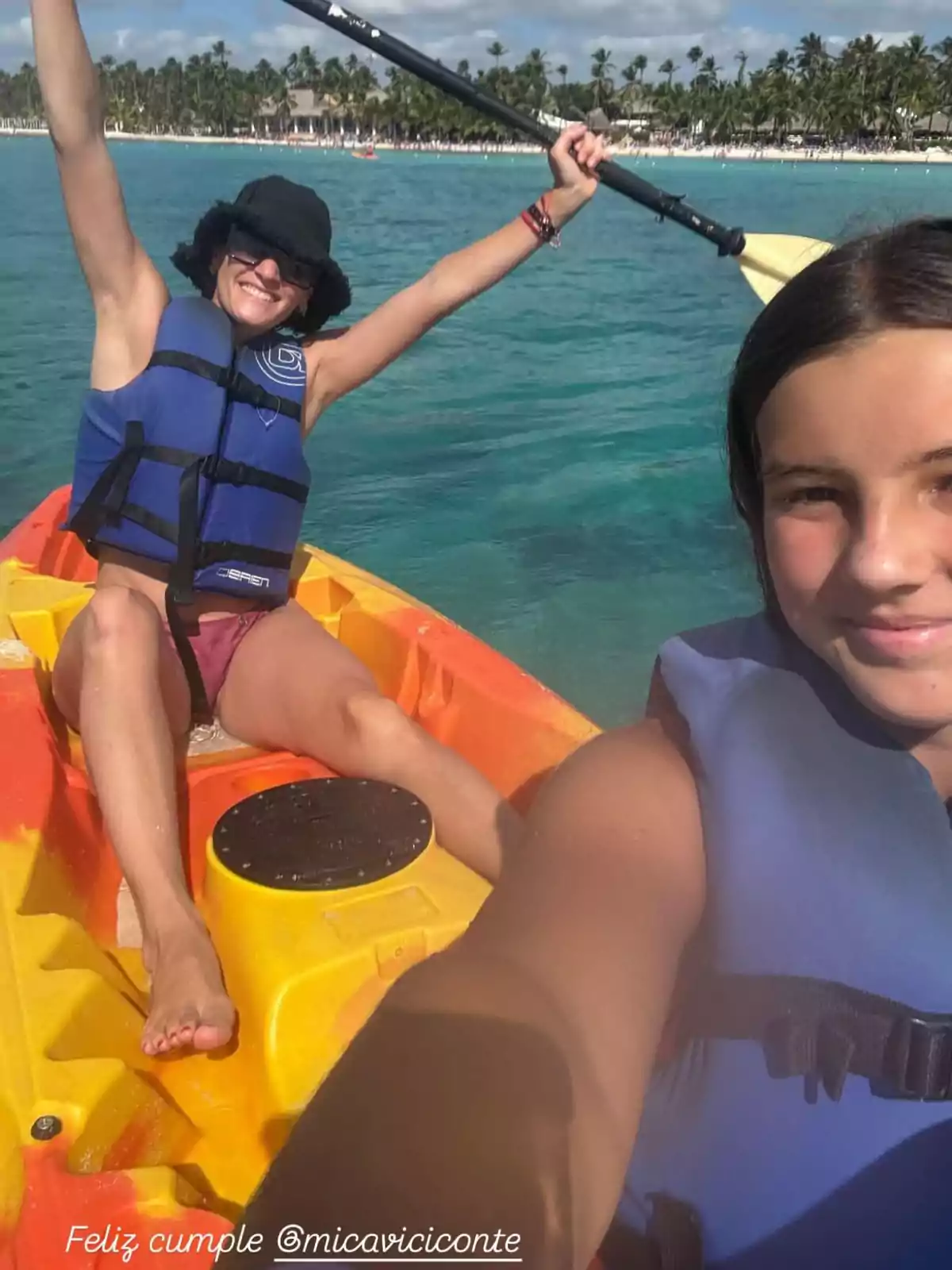 Two people enjoy a kayak ride in crystal-clear waters, one of them enthusiastically raises the paddle while the other takes a selfie, both wearing blue life jackets with palm trees in the background. Two people enjoy a kayak ride in crystal-clear waters, one of them enthusiastically raises the paddle while the other takes a selfie, both wearing blue life jackets with palm trees in the background.
