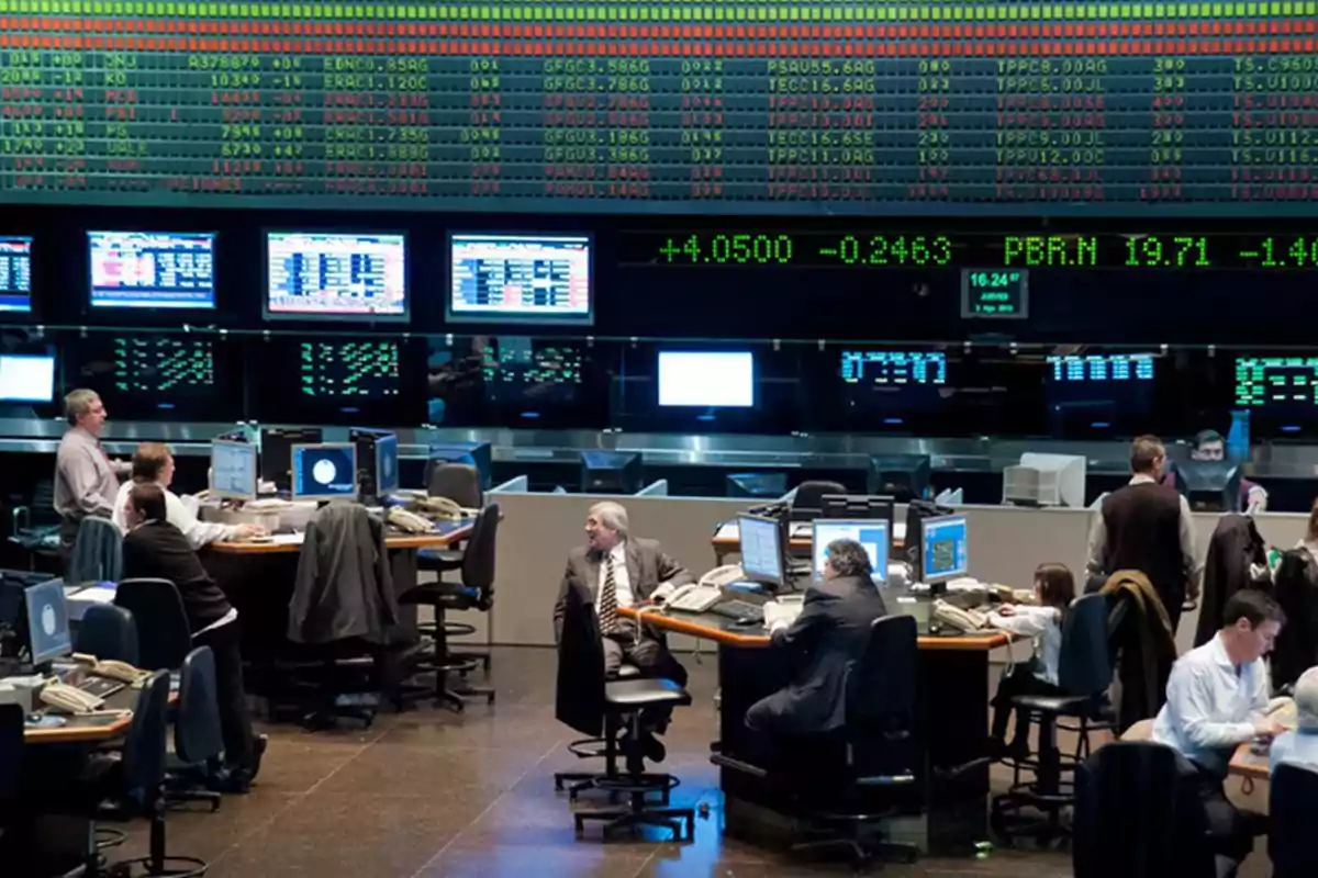 Personas trabajando en una sala de operaciones bursátiles con múltiples pantallas mostrando datos financieros.