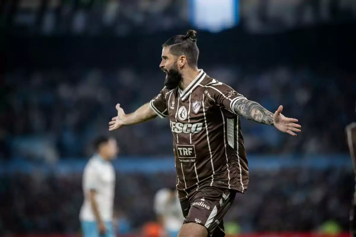 Un jugador de fútbol con barba y tatuajes celebra con los brazos abiertos en el campo mientras viste un uniforme marrón. Un jugador de fútbol con barba y tatuajes celebra con los brazos abiertos en el campo mientras viste un uniforme marrón.