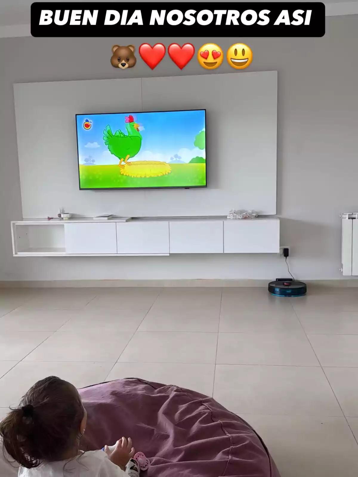 A girl sitting on a bean bag watches a TV showing a cartoon of a green chicken in a colorful setting, while a robot vacuum is on the floor. A girl sitting on a bean bag watches a TV showing a cartoon of a green chicken in a colorful setting, while a robot vacuum is on the floor.