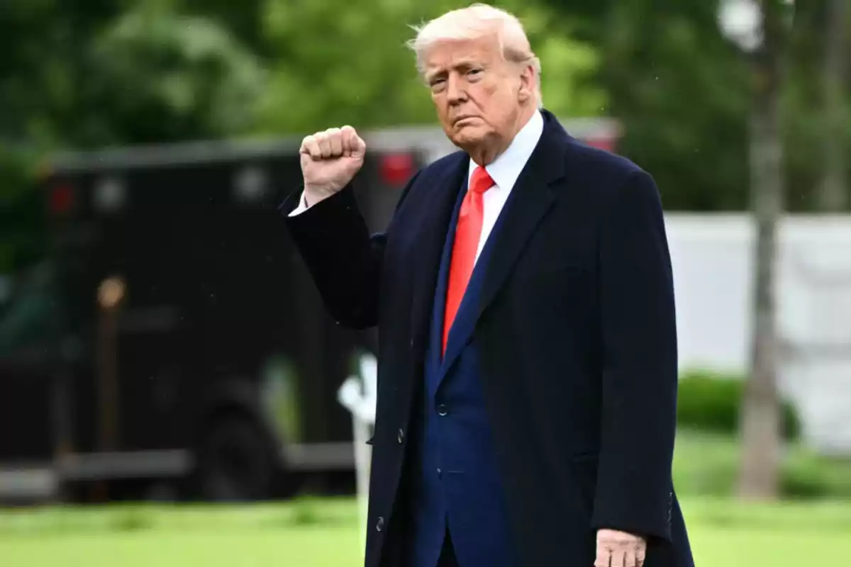 Hombre de cabello rubio con traje oscuro y corbata roja levantando el puño en un entorno al aire libre Hombre de cabello rubio con traje oscuro y corbata roja levantando el puño en un entorno al aire libre
