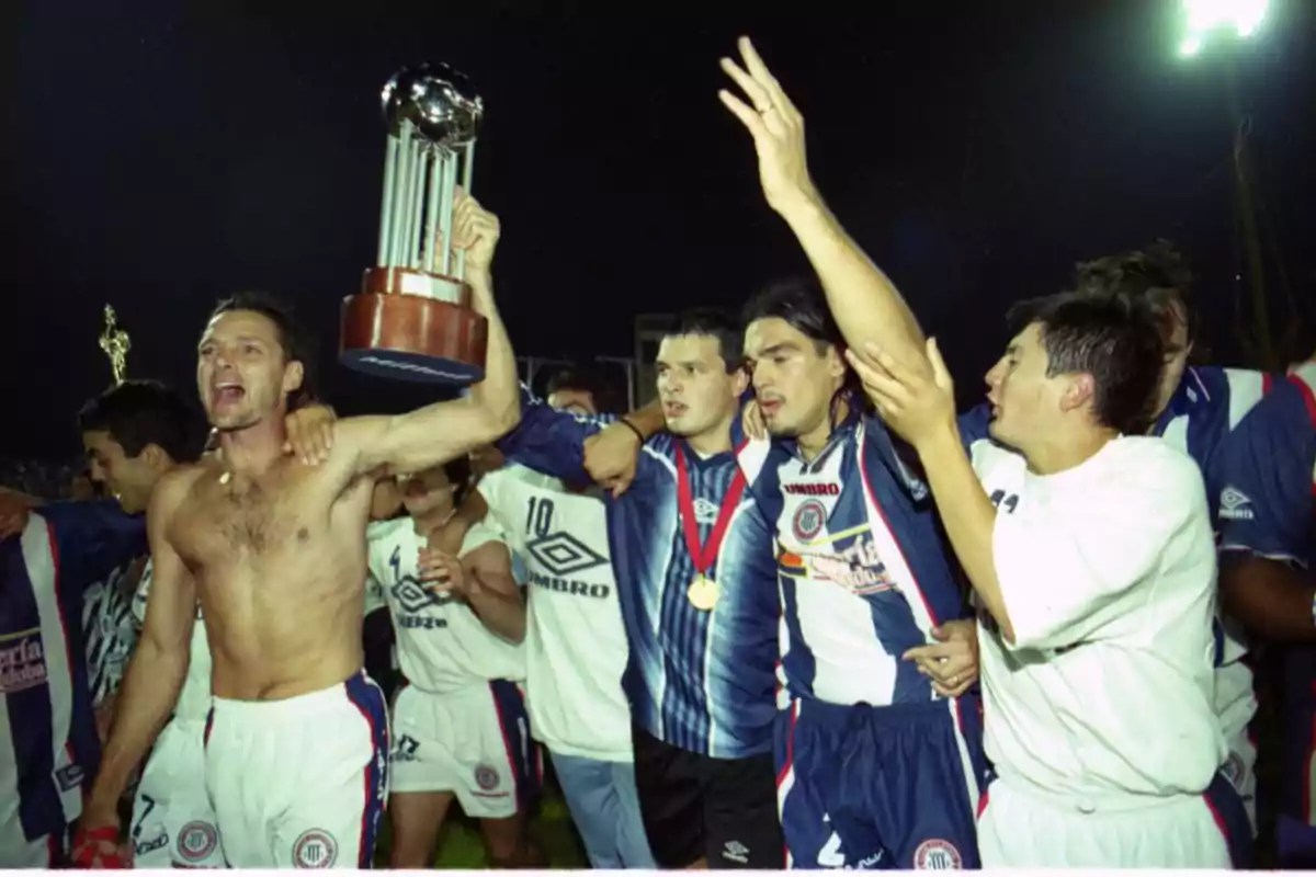 A group of players celebrates with a trophy held high, showing excitement and joy in a brightly lit stadium.