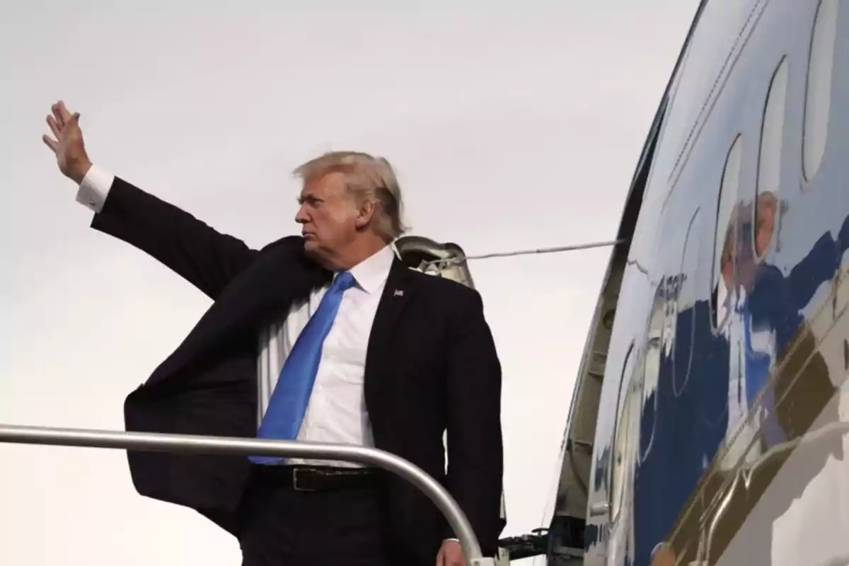 Hombre de traje oscuro y corbata azul saludando con la mano mientras sube las escaleras de un avión