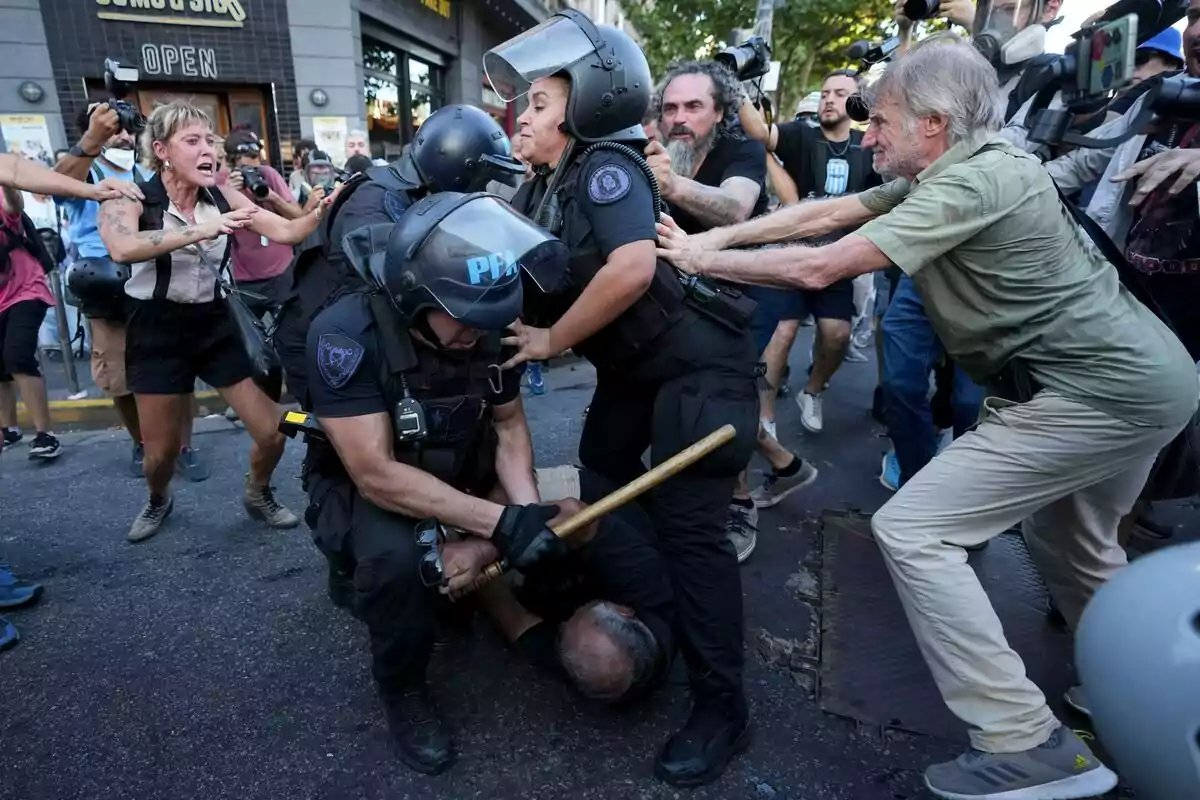 Una escena de protesta donde policías con equipo antidisturbios detienen a una persona en el suelo mientras otros manifestantes y fotógrafos los rodean.