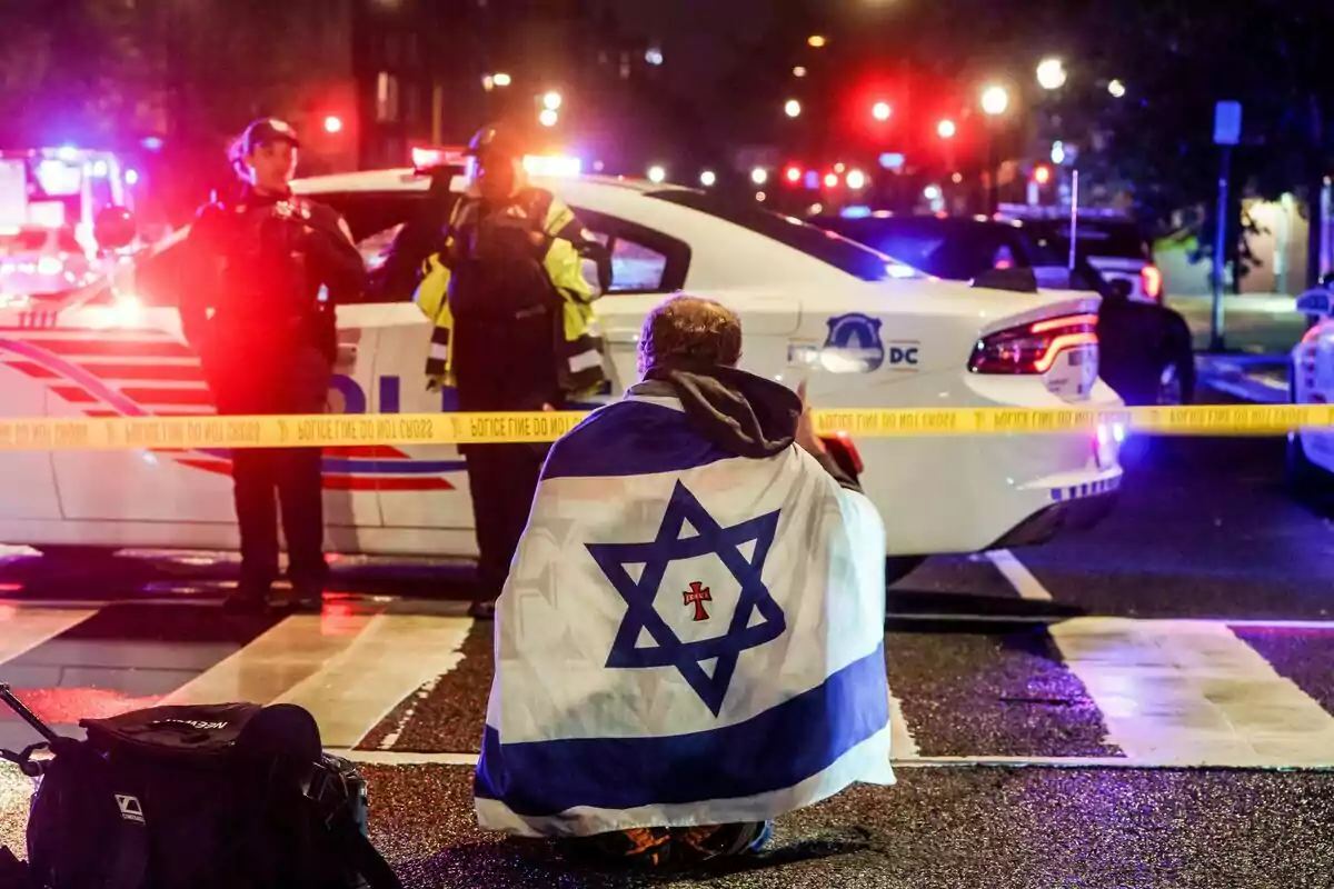 A person wrapped in a flag with the Star of David is sitting on the street in front of a police car and two officers, with emergency lights and caution tape at the scene. A person wrapped in a flag with the Star of David is sitting on the street in front of a police car and two officers, with emergency lights and caution tape at the scene.