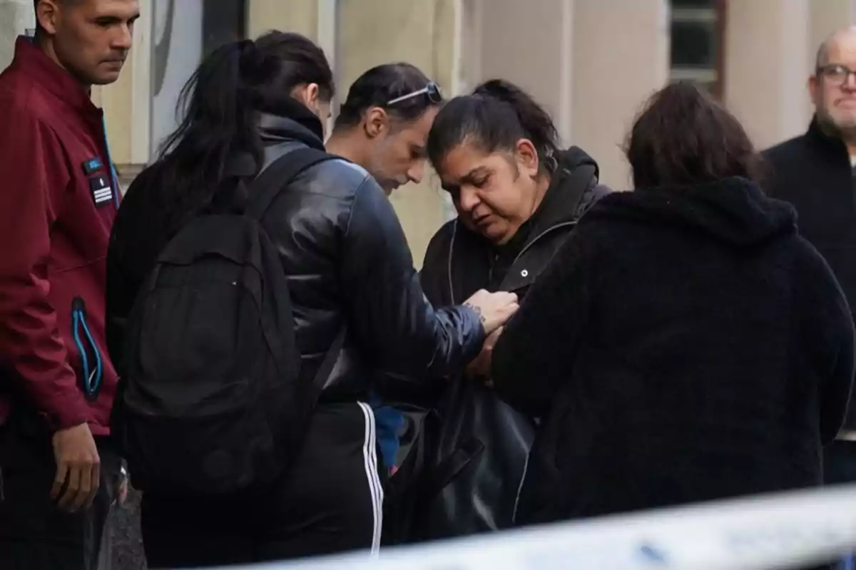 Un grupo de personas se encuentra reunido en la calle, algunas parecen estar consolándose mutuamente mientras un hombre observa la escena. Un grupo de personas se encuentra reunido en la calle, algunas parecen estar consolándose mutuamente mientras un hombre observa la escena.