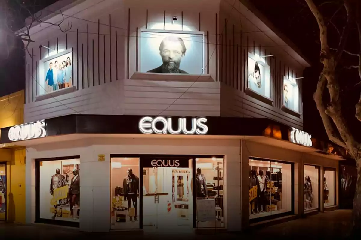 Facade of an Equus clothing store illuminated at night with mannequins in the display windows and posters of people on the upper part of the building