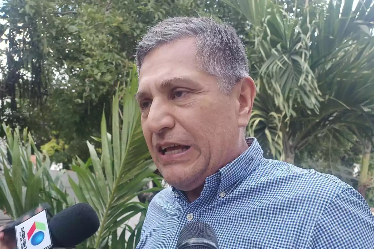 Gray-haired man in a blue plaid jersey speaking in front of microphones outdoors with plants in the background Gray-haired man in a blue plaid jersey speaking in front of microphones outdoors with plants in the background