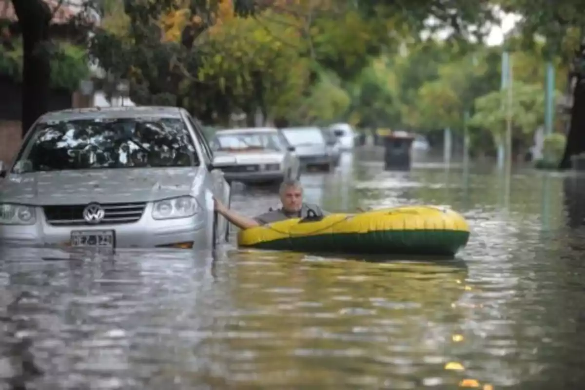 A man on an inflatable raft moves down a flooded street next to a partially submerged car.