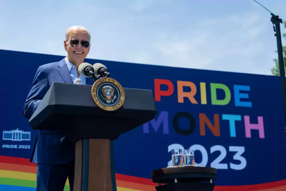 Un hombre con gafas de sol y traje azul habla en un podio con el sello presidencial de Estados Unidos, frente a un cartel que celebra el Mes del Orgullo 2023. Un hombre con gafas de sol y traje azul habla en un podio con el sello presidencial de Estados Unidos, frente a un cartel que celebra el Mes del Orgullo 2023.