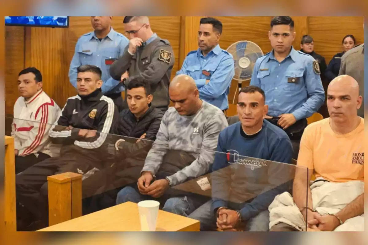Several men sitting in a courtroom with handcuffs on their hands and police officers standing behind them. Several men sitting in a courtroom with handcuffs on their hands and police officers standing behind them.