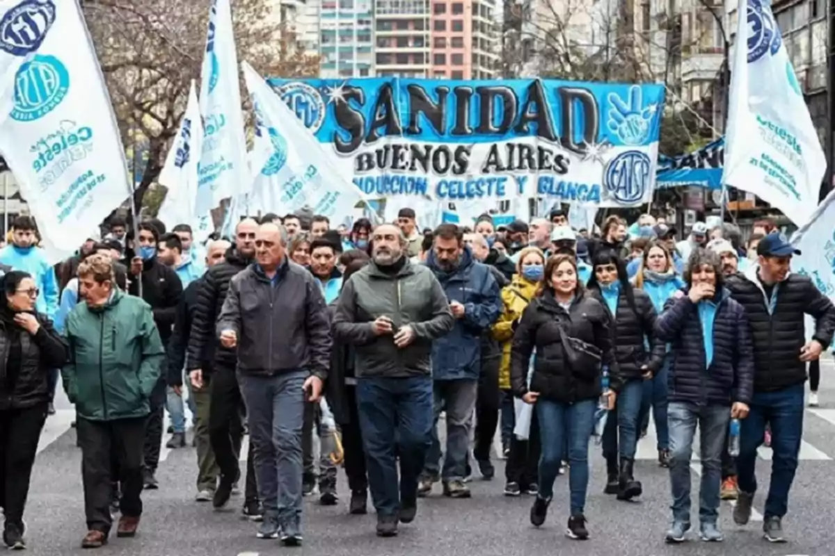 Un grupo de personas marcha por una calle sosteniendo banderas y un cartel grande que dice Sanidad Buenos Aires, rodeados de edificios y árboles. Un grupo de personas marcha por una calle sosteniendo banderas y un cartel grande que dice Sanidad Buenos Aires, rodeados de edificios y árboles.