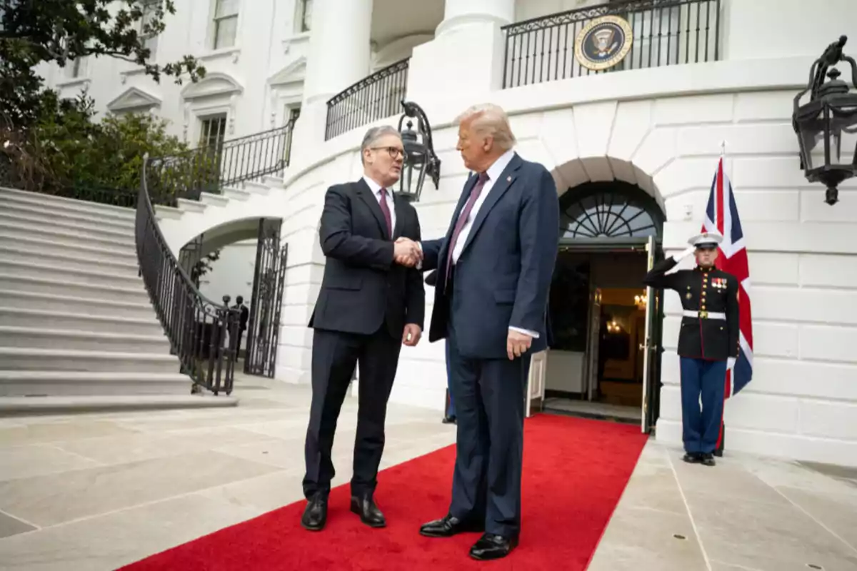 Two men in suits shake hands on a red carpet in front of a white building with columns and a guard in the background. Two men in suits shake hands on a red carpet in front of a white building with columns and a guard in the background.