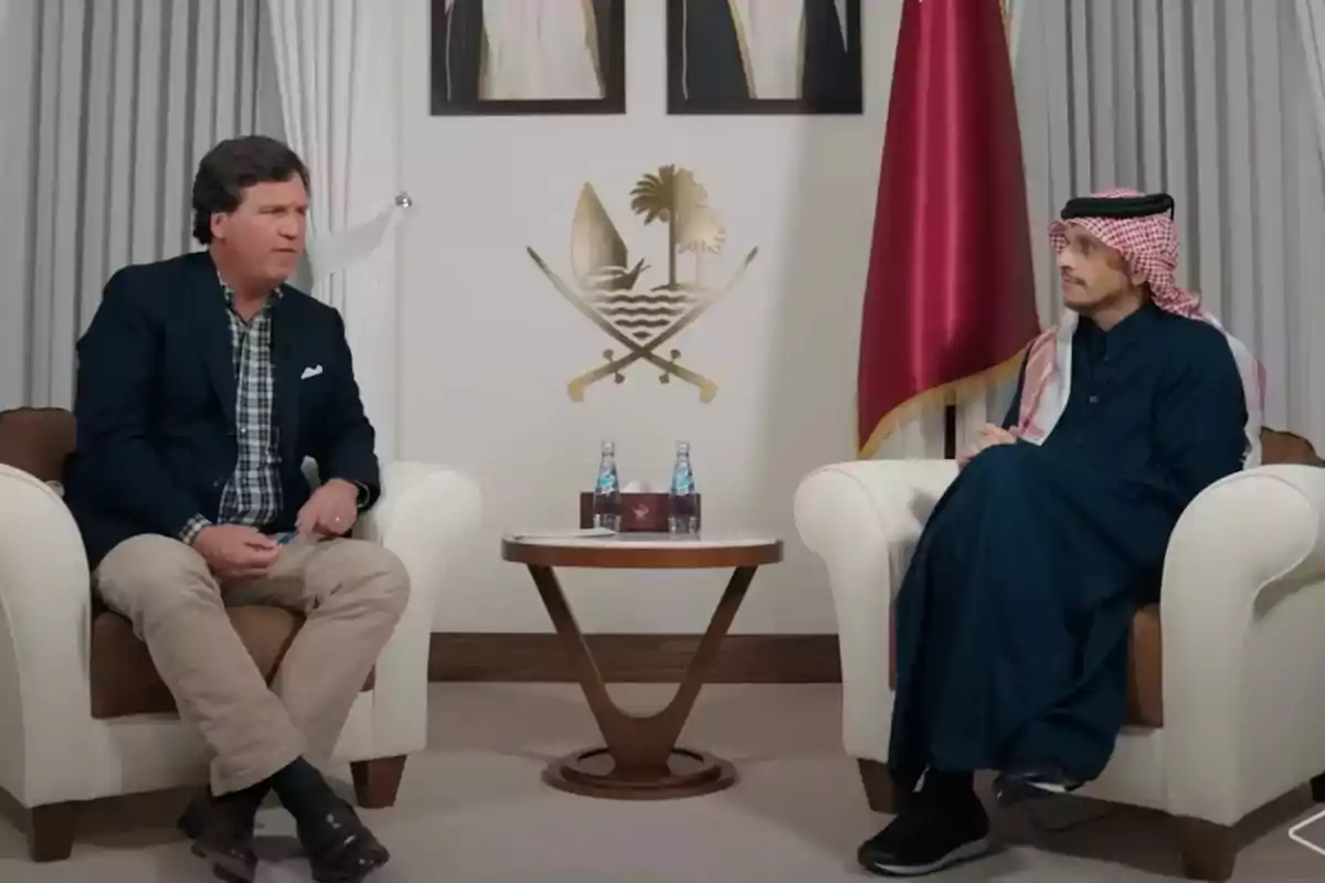 Two men sitting in white armchairs are talking in an elegant room with a small table between them, on which there are two bottles of water, a box of tissues, and a red flag in the background.