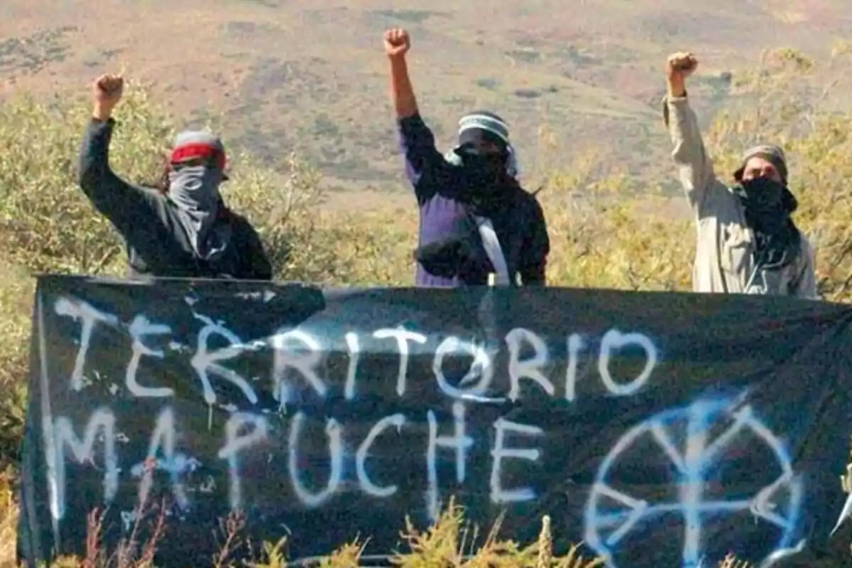 Tres personas con el rostro cubierto levantan el puño detrás de una pancarta que dice "Territorio Mapuche" en un paisaje natural.