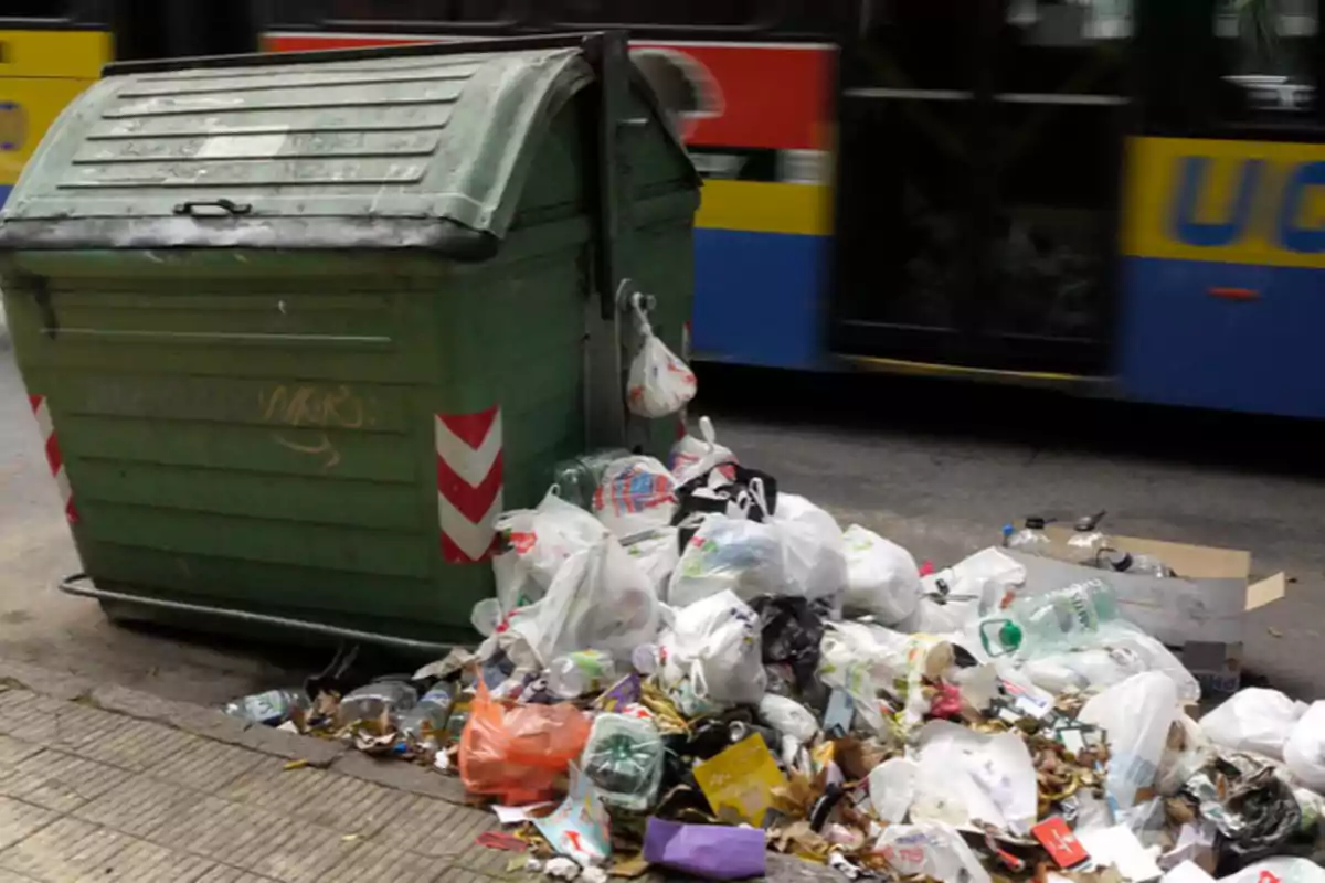 Contenedor de basura verde con bolsas de basura acumuladas alrededor en una acera junto a un autobús. Contenedor de basura verde con bolsas de basura acumuladas alrededor en una acera junto a un autobús.