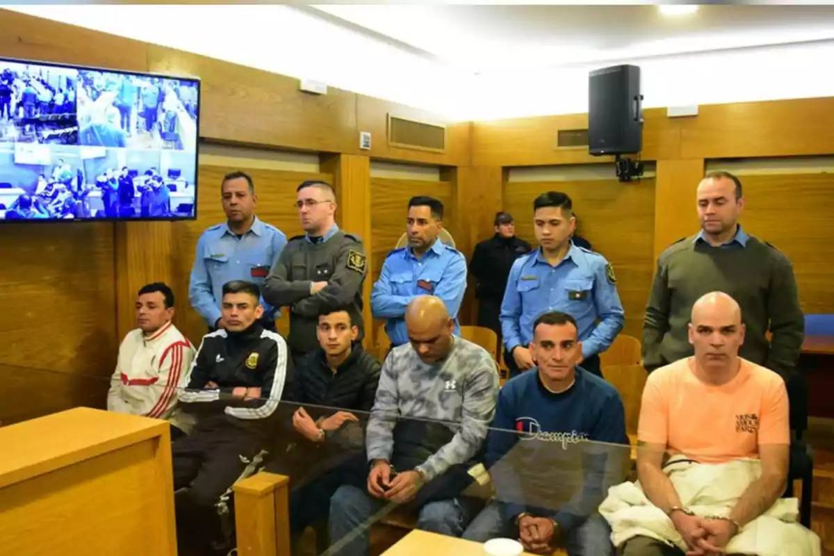 Several men seated in a courtroom with police officers standing behind them and a screen displaying security footage in the background