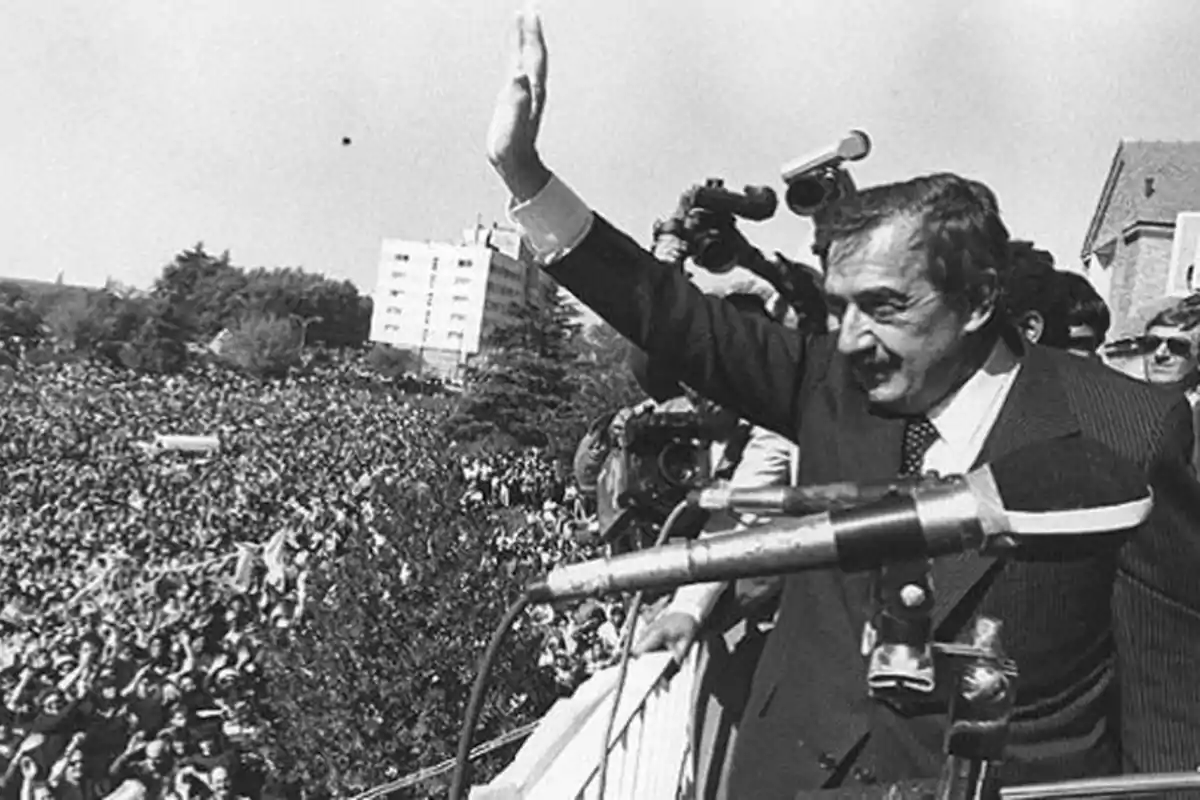 A man in a suit waves to a crowd from a balcony while being photographed and filmed by several cameras.