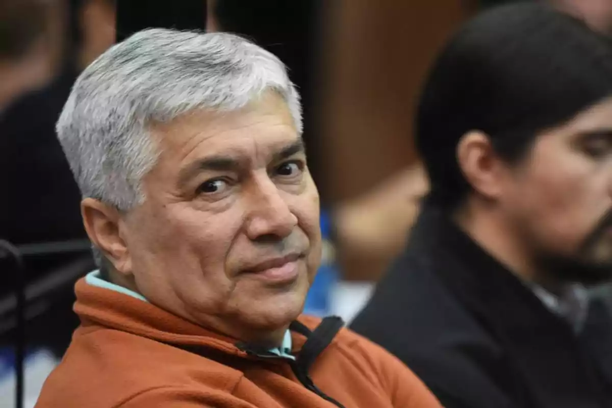 Hombre de cabello canoso y suéter marrón mira hacia la cámara mientras está sentado en un lugar público Hombre de cabello canoso y suéter marrón mira hacia la cámara mientras está sentado en un lugar público
