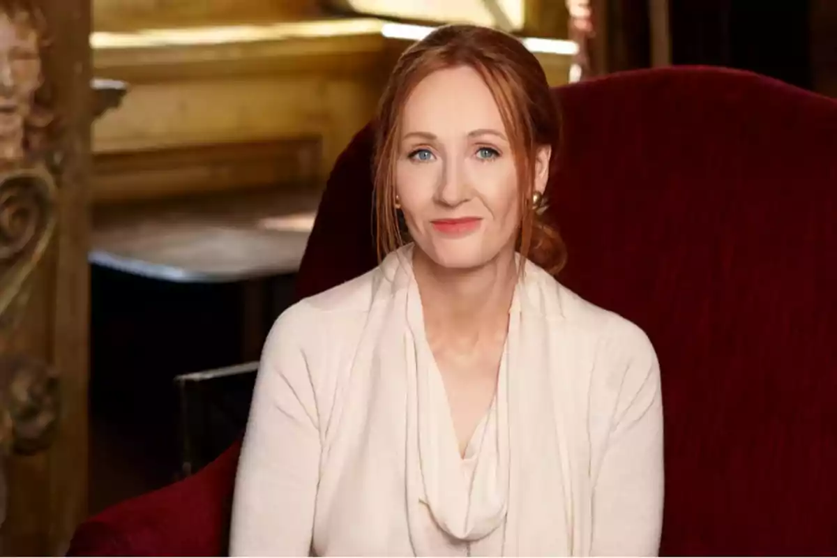 Red-haired woman with light eyes sitting on a red armchair, wearing a beige sweater and smiling slightly in an elegant setting.