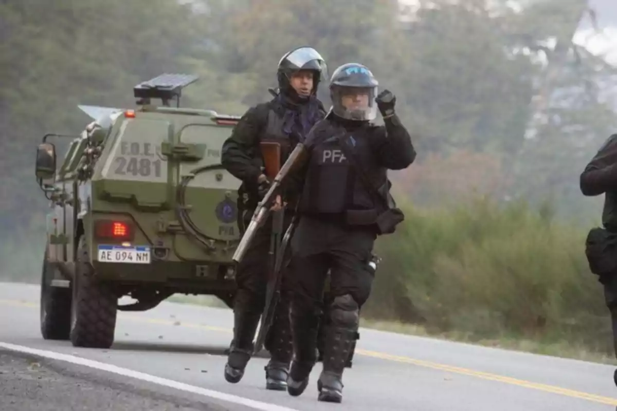 Policías con equipo antidisturbios caminando junto a un vehículo blindado en una carretera.