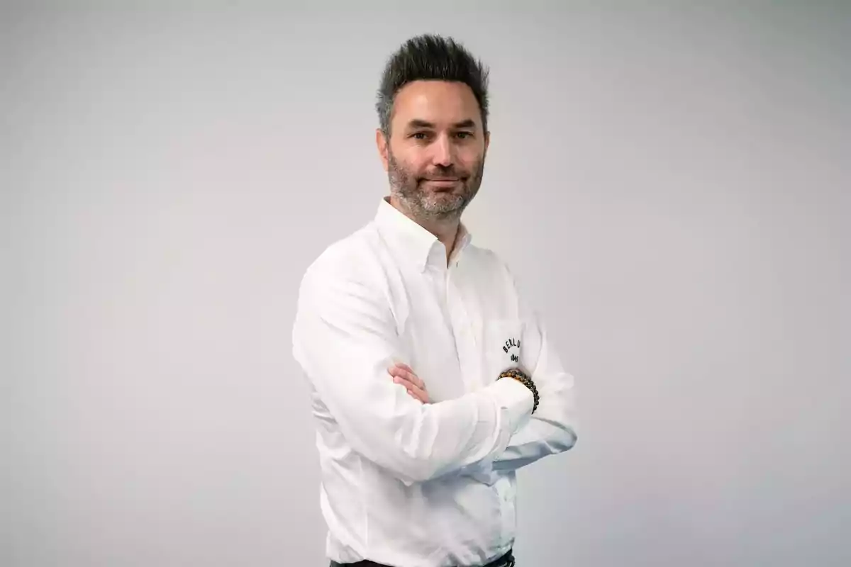 Hombre de cabello oscuro y barba con camisa blanca de manga larga posando con los brazos cruzados frente a un fondo gris claro Hombre de cabello oscuro y barba con camisa blanca de manga larga posando con los brazos cruzados frente a un fondo gris claro
