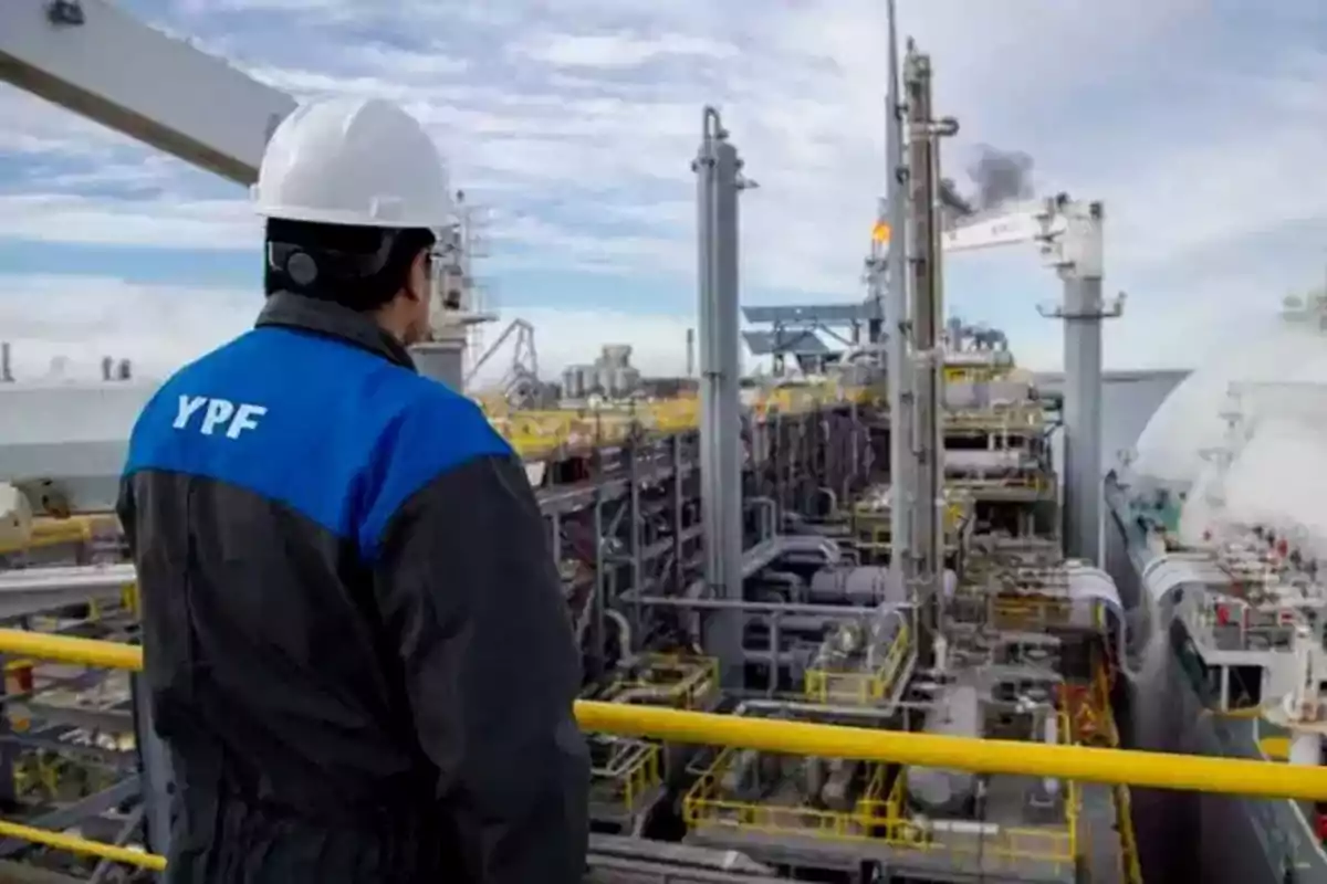 Un trabajador con casco y chaqueta de YPF observa una planta industrial con estructuras metálicas y tuberías.