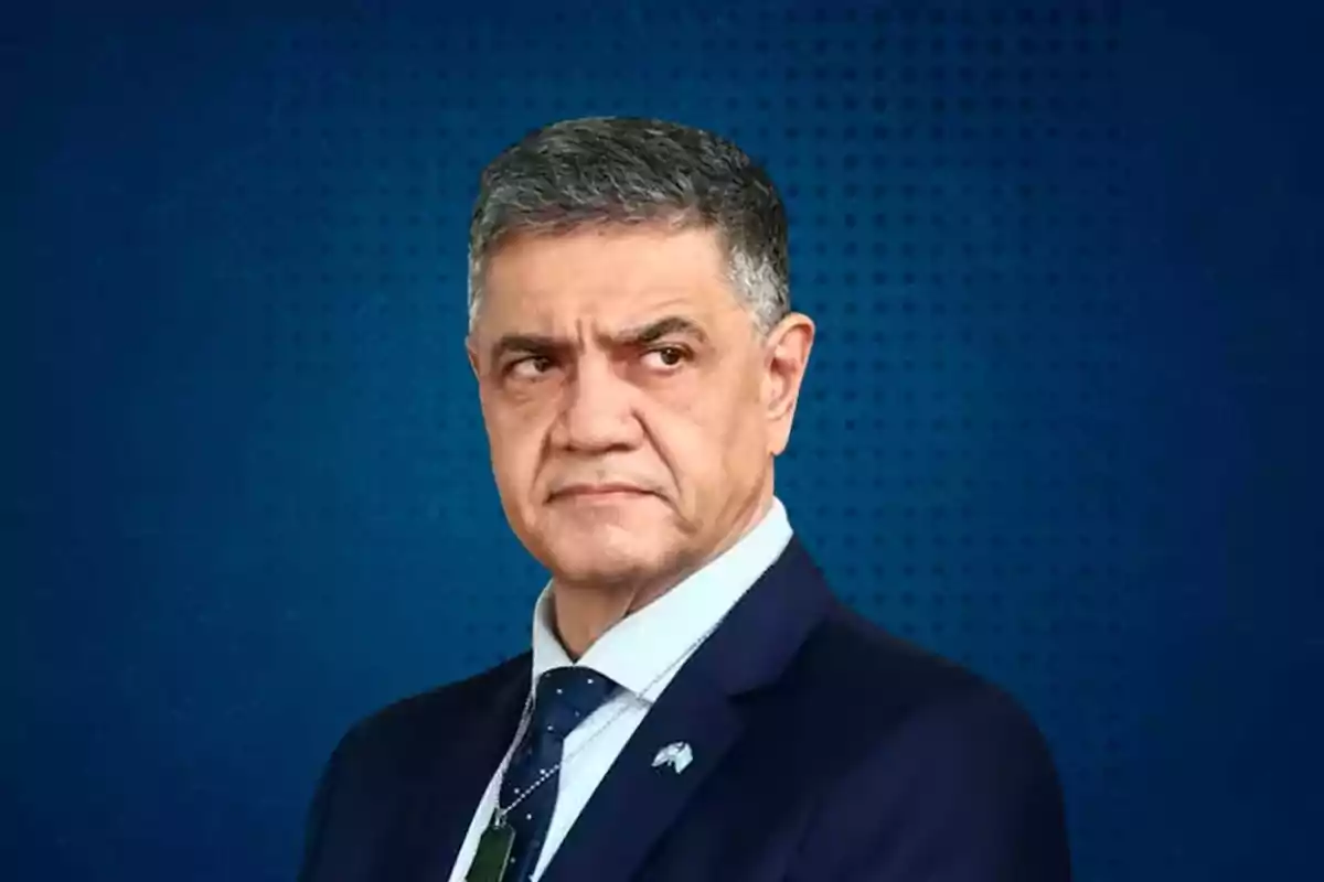 A gray-haired man with a serious expression wears a dark suit with a tie against a blue background.