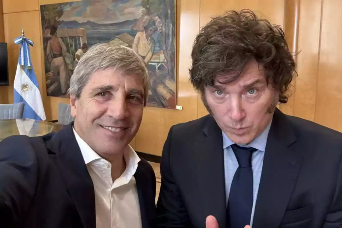 Two men pose for a selfie in an office with an Argentine flag and a painting on the wall.