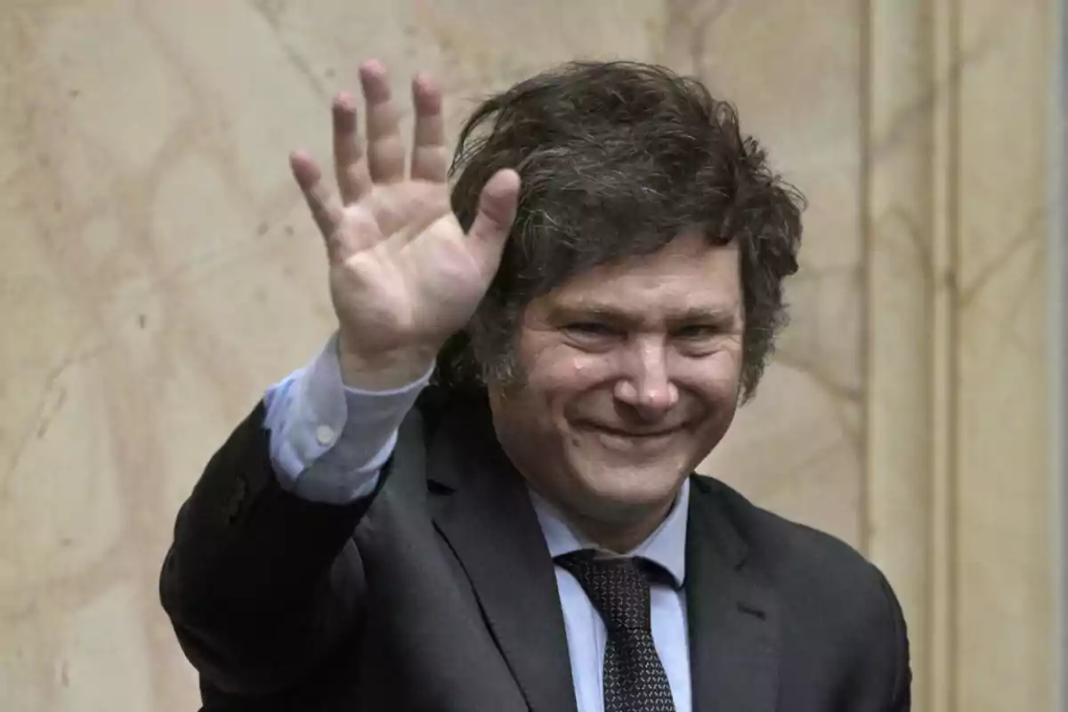 Hombre con traje oscuro y corbata, sonriendo y saludando con la mano levantada.