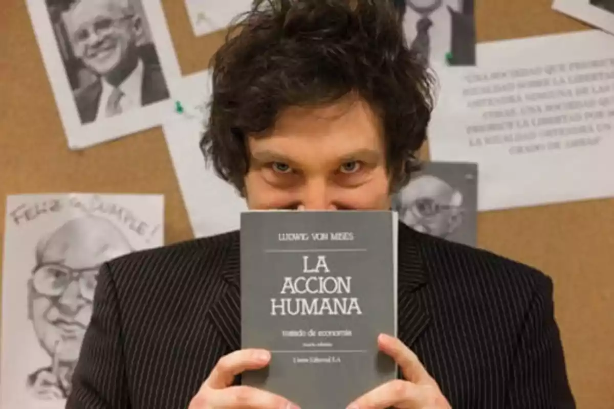 Hombre de cabello oscuro y rizado sostiene un libro titulado La Acción Humana frente a su rostro mientras mira a la cámara, con un fondo de imágenes y dibujos en una pared de corcho