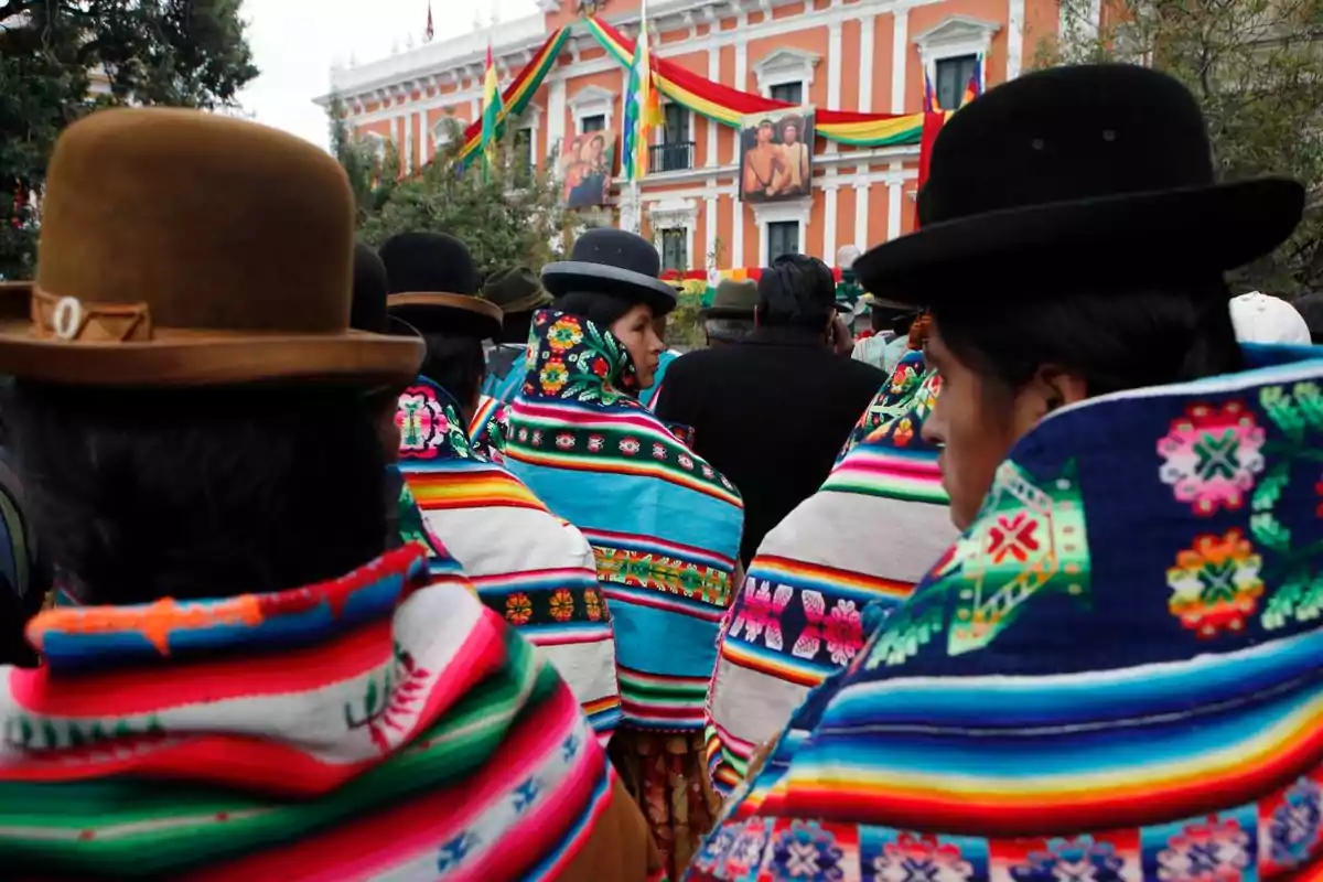 Women wearing traditional hats and colorful blankets participate in an event in front of a building decorated with flags and portraits in Bolivia. Women wearing traditional hats and colorful blankets participate in an event in front of a building decorated with flags and portraits in Bolivia.