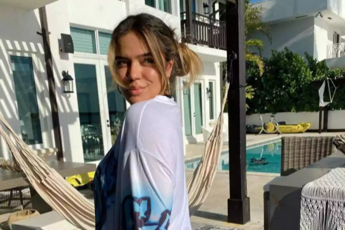 Joven con cabello recogido y camiseta blanca posando sonriente en un patio con piscina, hamaca y muebles de exterior en un día soleado Joven con cabello recogido y camiseta blanca posando sonriente en un patio con piscina, hamaca y muebles de exterior en un día soleado