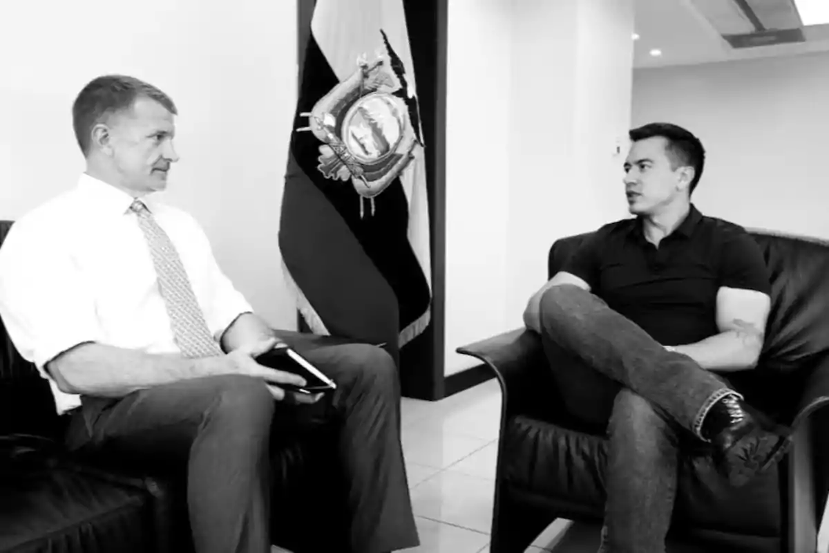 Two men sitting in armchairs are talking in an office with a flag in the background.