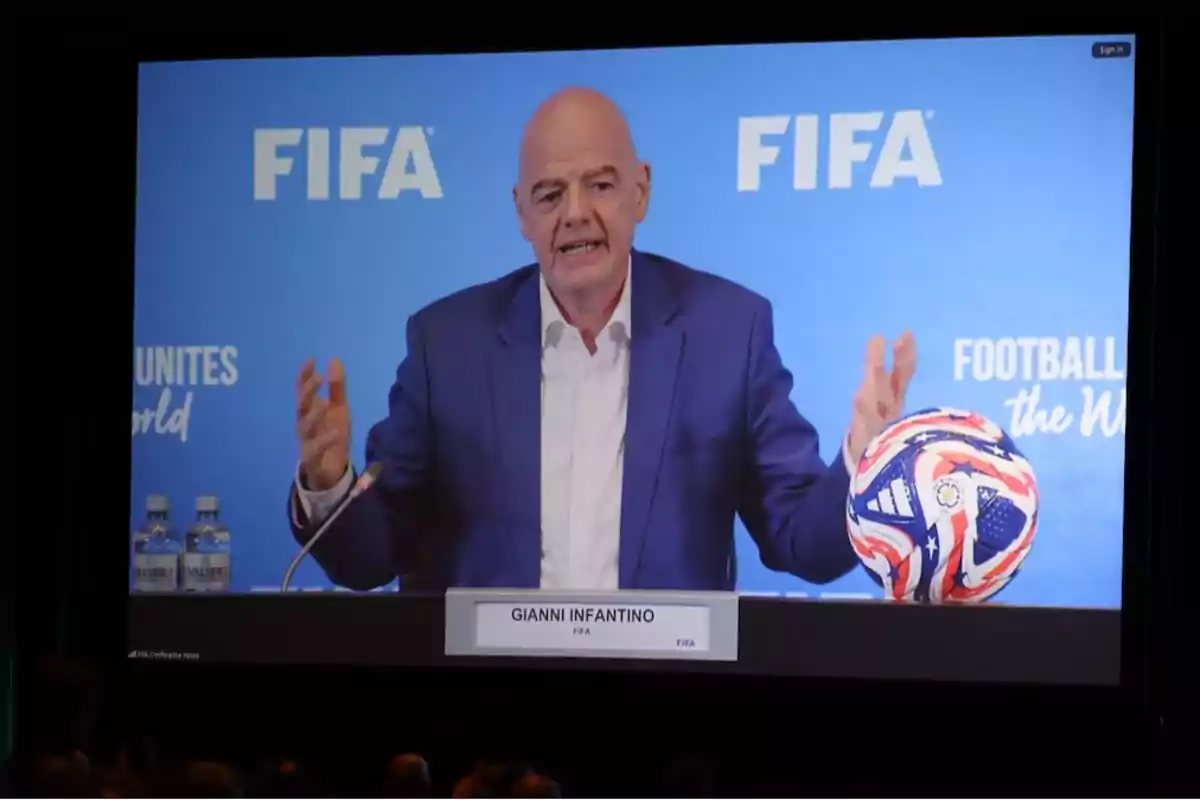 A man at a FIFA press conference, sitting in front of a microphone with a soccer ball next to him.