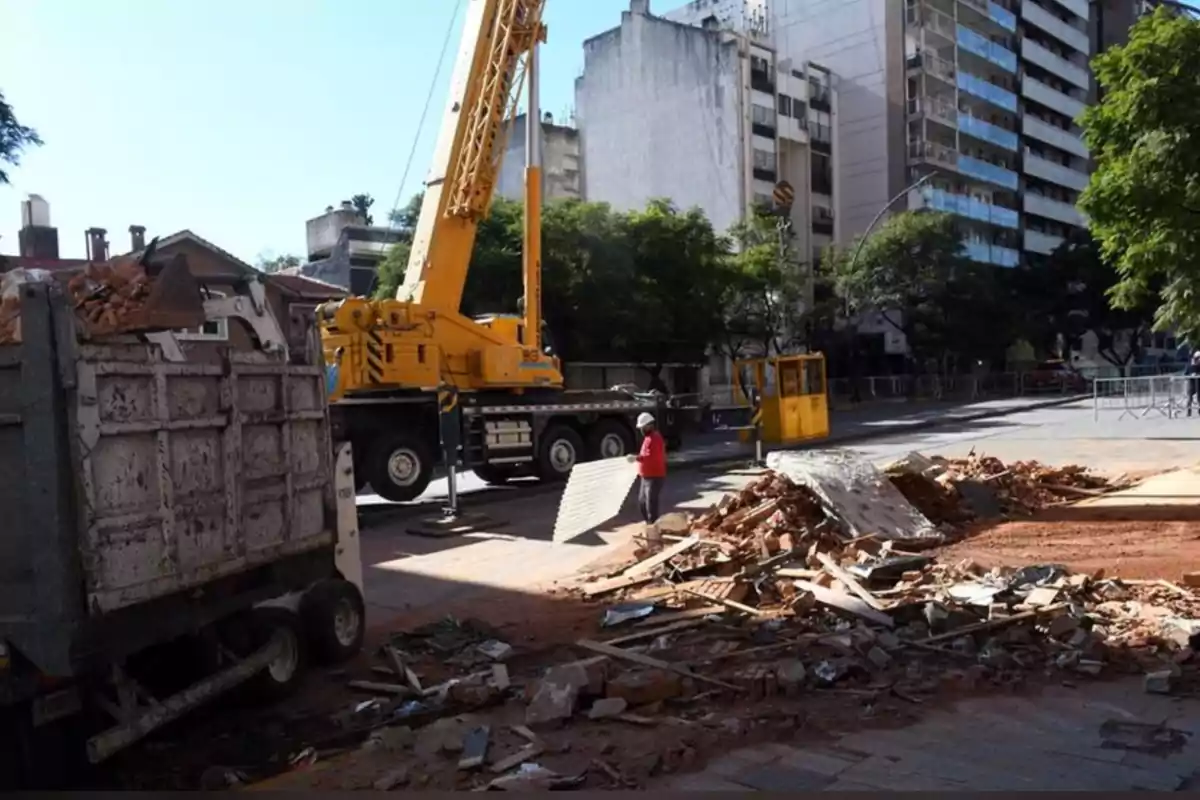 Un camión y una grúa trabajan en la remoción de escombros en una calle urbana mientras una persona con casco y chaleco manipula una plancha metálica Un camión y una grúa trabajan en la remoción de escombros en una calle urbana mientras una persona con casco y chaleco manipula una plancha metálica