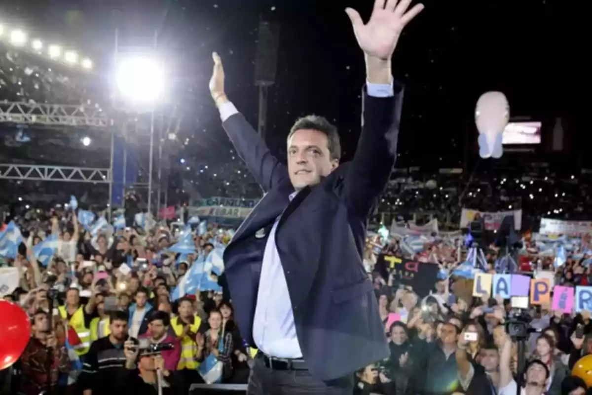 Persona levantando los brazos en un mitin político con una multitud de personas y banderas en el fondo.