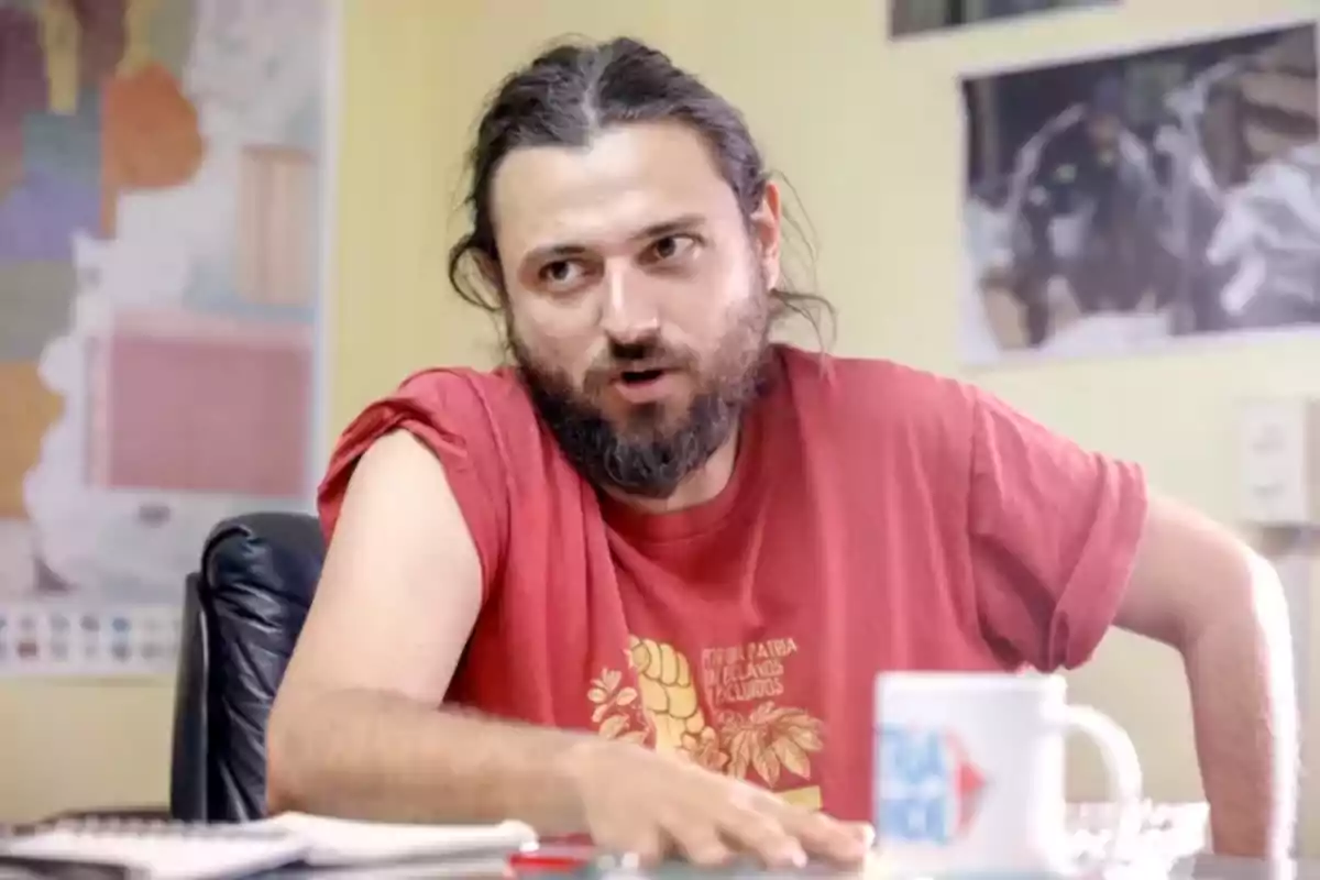 Un hombre con barba y cabello largo está sentado en una oficina, vistiendo una camiseta roja y hablando mientras gesticula con la mano.