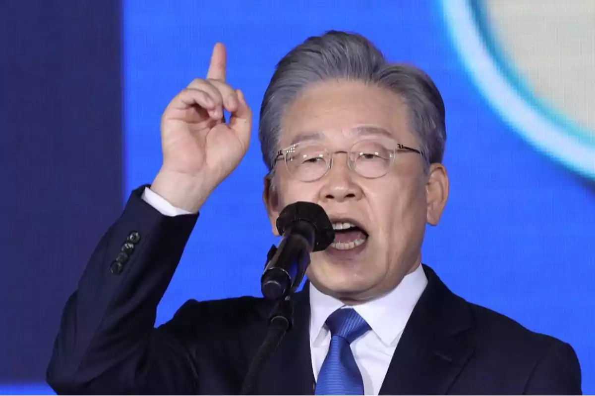 Hombre de cabello canoso y gafas hablando en un micrófono mientras levanta un dedo, vistiendo traje azul y corbata, con fondo azul
