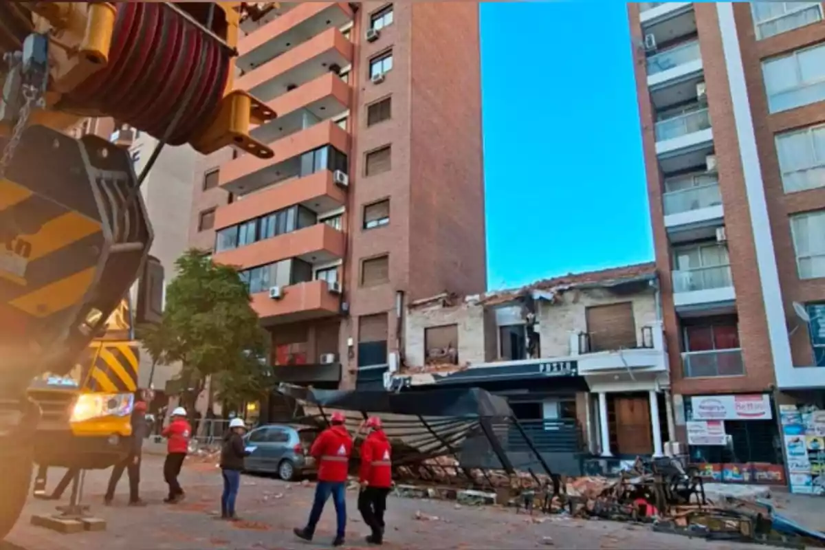 Varias personas con cascos y chalecos observan los daños causados por el derrumbe parcial de un edificio en una calle urbana, con escombros y una estructura metálica caída frente a la fachada afectada.