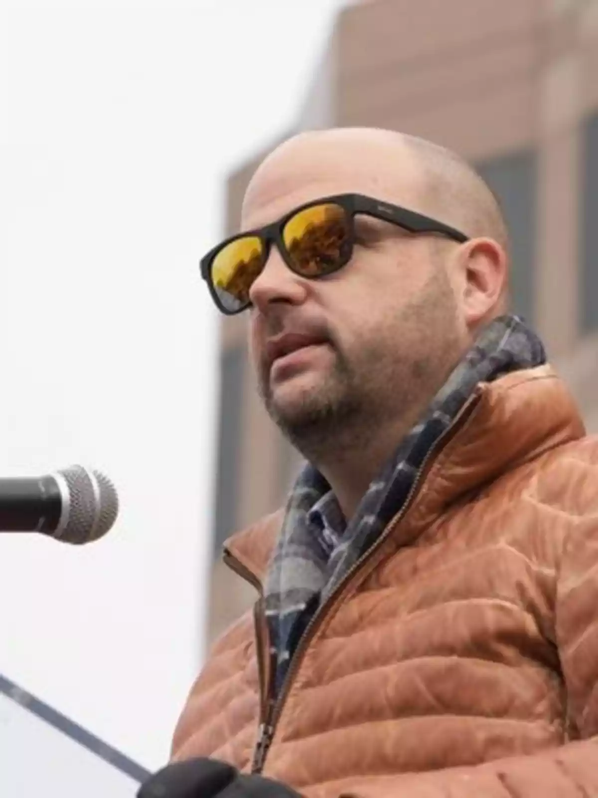 Man in a brown jacket and scarf, wearing sunglasses, speaking in front of a microphone. Man in a brown jacket and scarf, wearing sunglasses, speaking in front of a microphone.